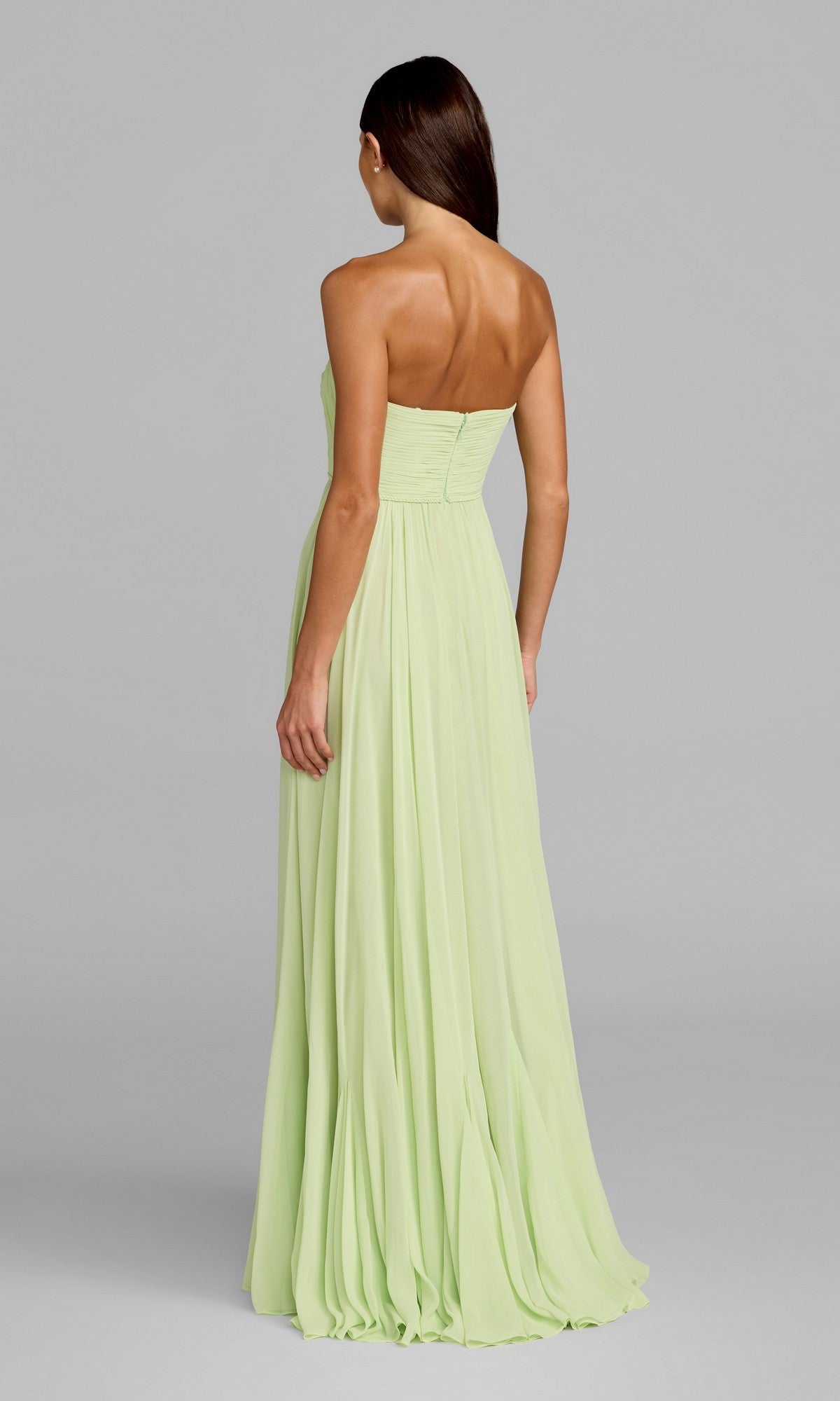 A woman with long brown hair is shown standing and facing away on a plain gray background, wearing the Mac Duggal 68847 long formal dress—a strapless chiffon gown featuring an elegant light green A-line skirt.